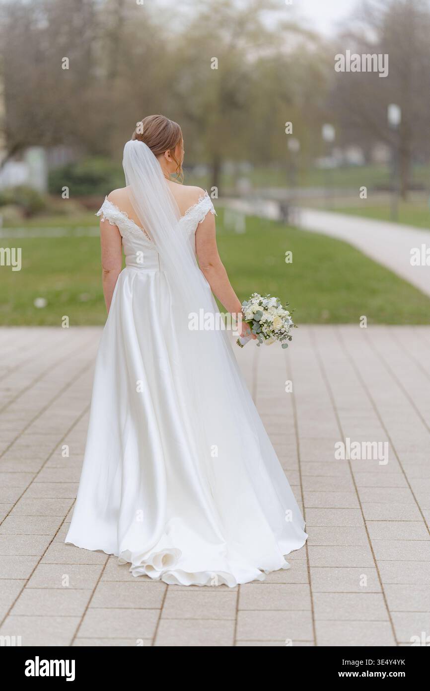 Sposa che cammina da sola all'aperto con un abito da sposa in possesso di bouquet. Momento romantico ed emotivo prima della cerimonia. Concetto di matrimonio elegante e naturale. Foto Stock