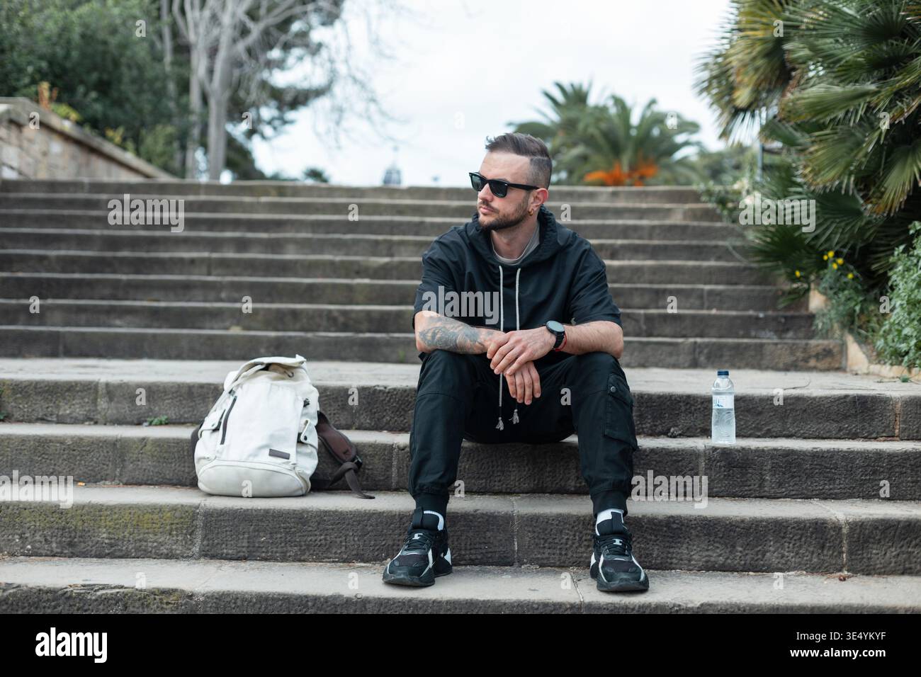 Elegante e affascinante viaggiatore di moda con occhiali da sole con cappuccio e scarpe da ginnastica sedute sui gradini con zaino nel parco di Barcellona Foto Stock