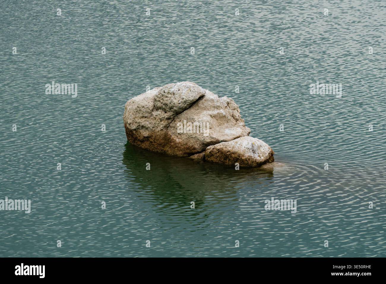 Un masso di colore chiaro si trova isolato nel mezzo dell'acqua ondulata profonda. La struttura della roccia è ruvida e intemprata. L'acqua è di colore grigio scuro Foto Stock