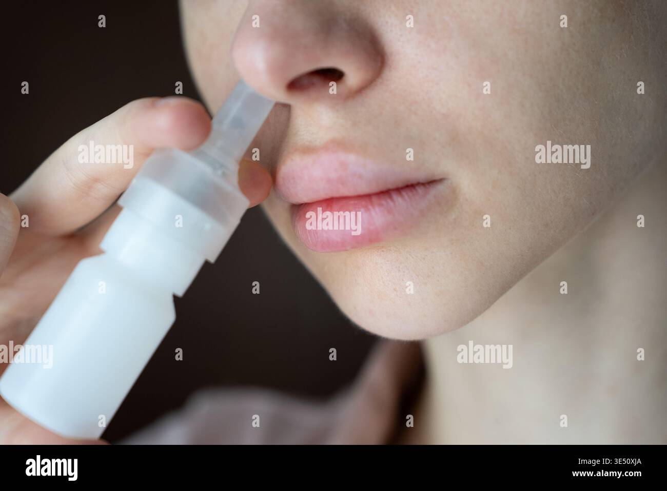 Una giovane donna applica uno spray nasale al naso per un sollievo, concentrandosi sulle sue caratteristiche delicate e sulla bottiglia medica in uno scatto ravvicinato e morbido. Foto Stock