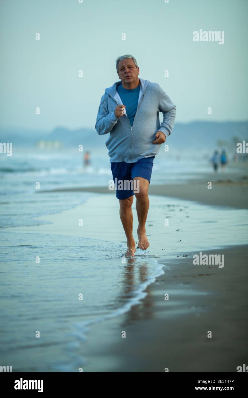 Uomo maturo che fa jogging a piedi nudi lungo il litorale in spiaggia, indossa una felpa con cappuccio grigia e pantaloncini blu, con onde dolci che gli si infrangono ai piedi e una nebulosa Foto Stock