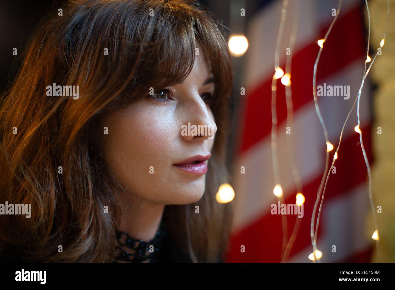 Modello femminile con lunghi capelli marroni posa davanti a una bandiera americana e luci decorative, mostrando una vista ravvicinata del profilo con illuminazione soffusa e. Foto Stock