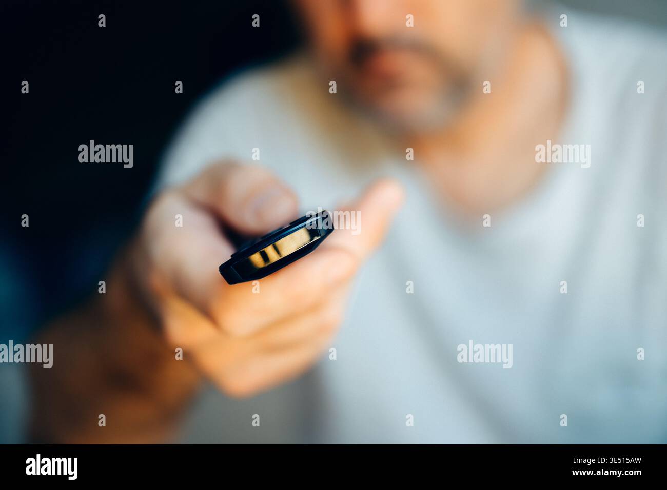 Primo piano di un uomo che tiene in mano il telecomando TV con messa a fuoco selettiva su tasti e dita, volto sfocato sullo sfondo scuro del salotto Foto Stock