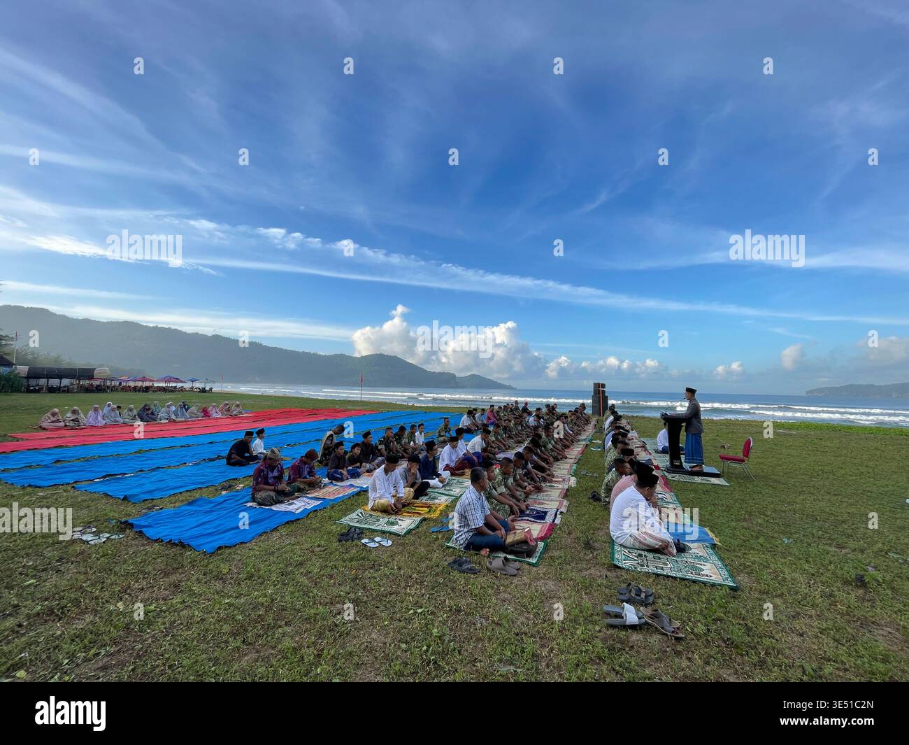 Uomini musulmani che eseguono le preghiere di Eid al-Fitr in righe ordinate su un campo verde vicino alla spiaggia. Una tranquilla cerimonia religiosa con tappeti verdi, uno sfondo - Immagine stock catturata con smartphone