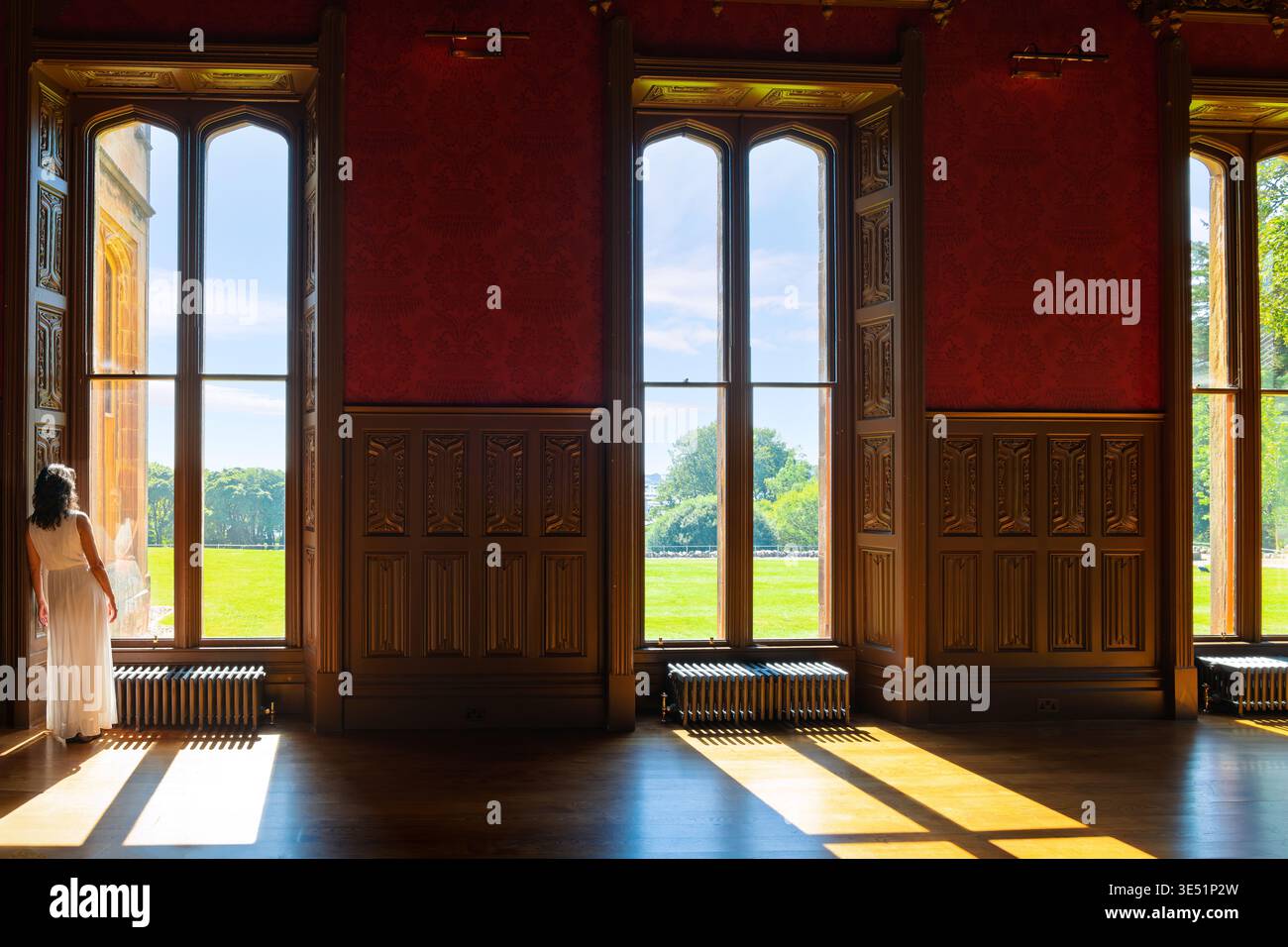 Una donna sta alla luce del sole guardando fuori da una finestra all'interno della sala del castello di Lews, affacciata su un prato e un giardino a Stornoway, in Scozia. Foto Stock