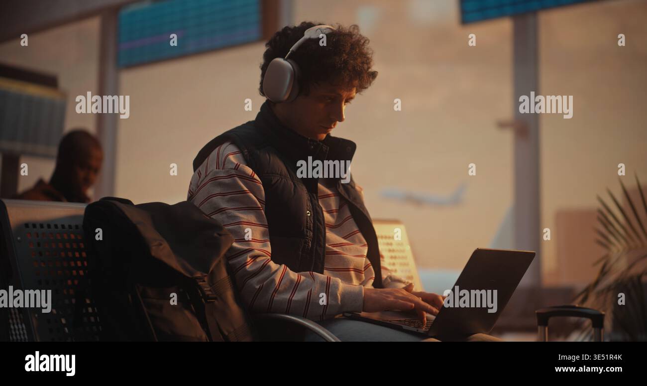 Young Man con capelli ricci siede vicino a un grande finestrino nella lounge partenze dell'aeroporto. Uomo focalizzato sullo schermo del notebook, dita che si muovono rapidamente sulla tastiera come scrive il codice. Produttività di Modern Programmer. Foto Stock