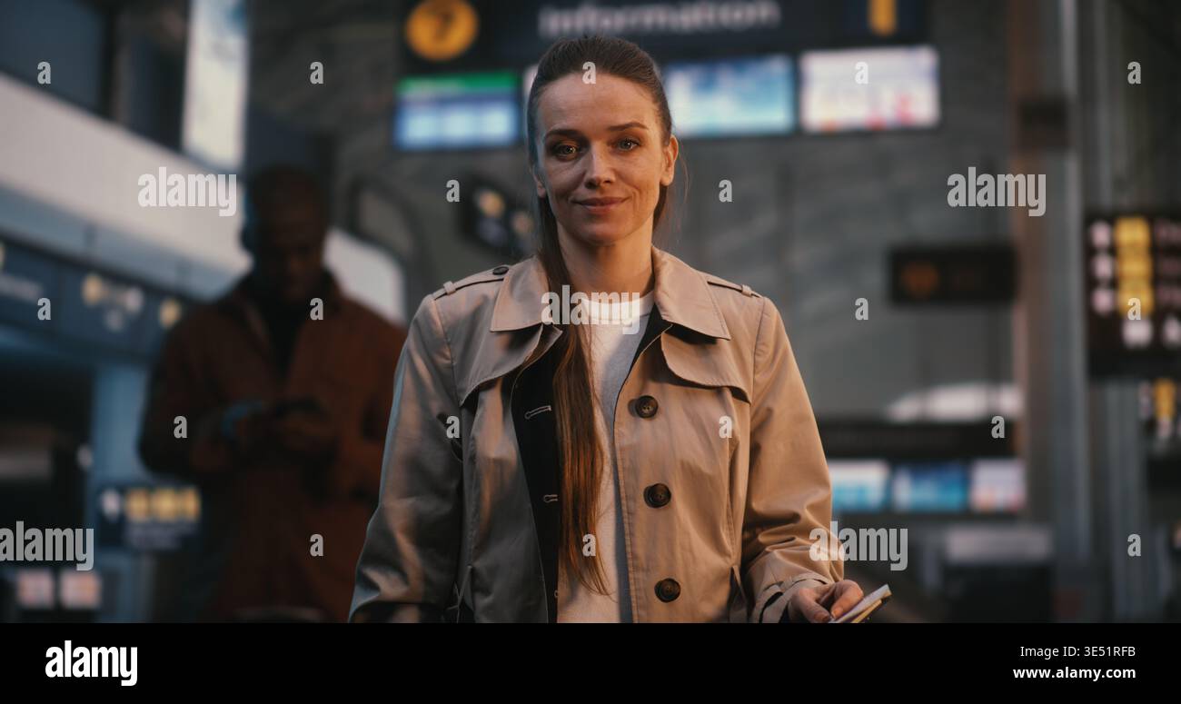 Il ritratto della splendida giovane donna con sorriso si trova nel vivace terminal dell'aeroporto. Vestita con un classico Trench beige femminile che esprime entusiasmo e gioia per il prossimo viaggio. Donna di stile. Foto Stock