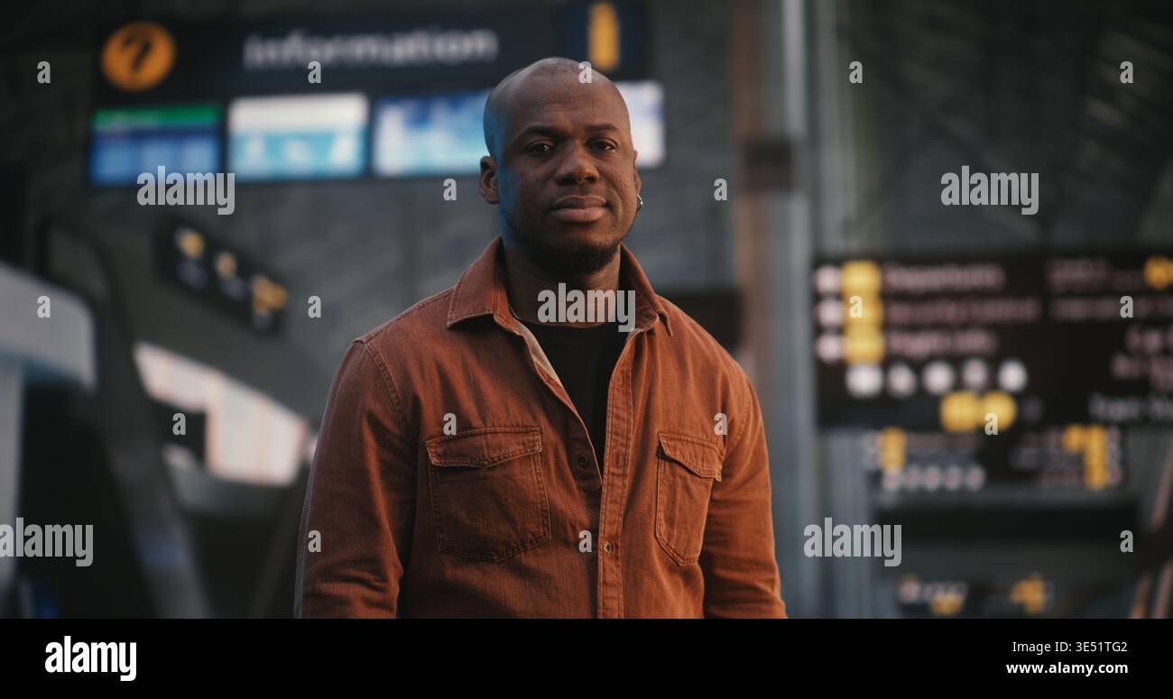 Maschio afroamericano si erge con sicurezza e sorriso sottile nell'area partenze dell'aeroporto internazionale. Terminal dell'ambiente vivace, con le persone che camminano, in evidenza il comportamento incentrato sull'uomo. Verticale. Foto Stock