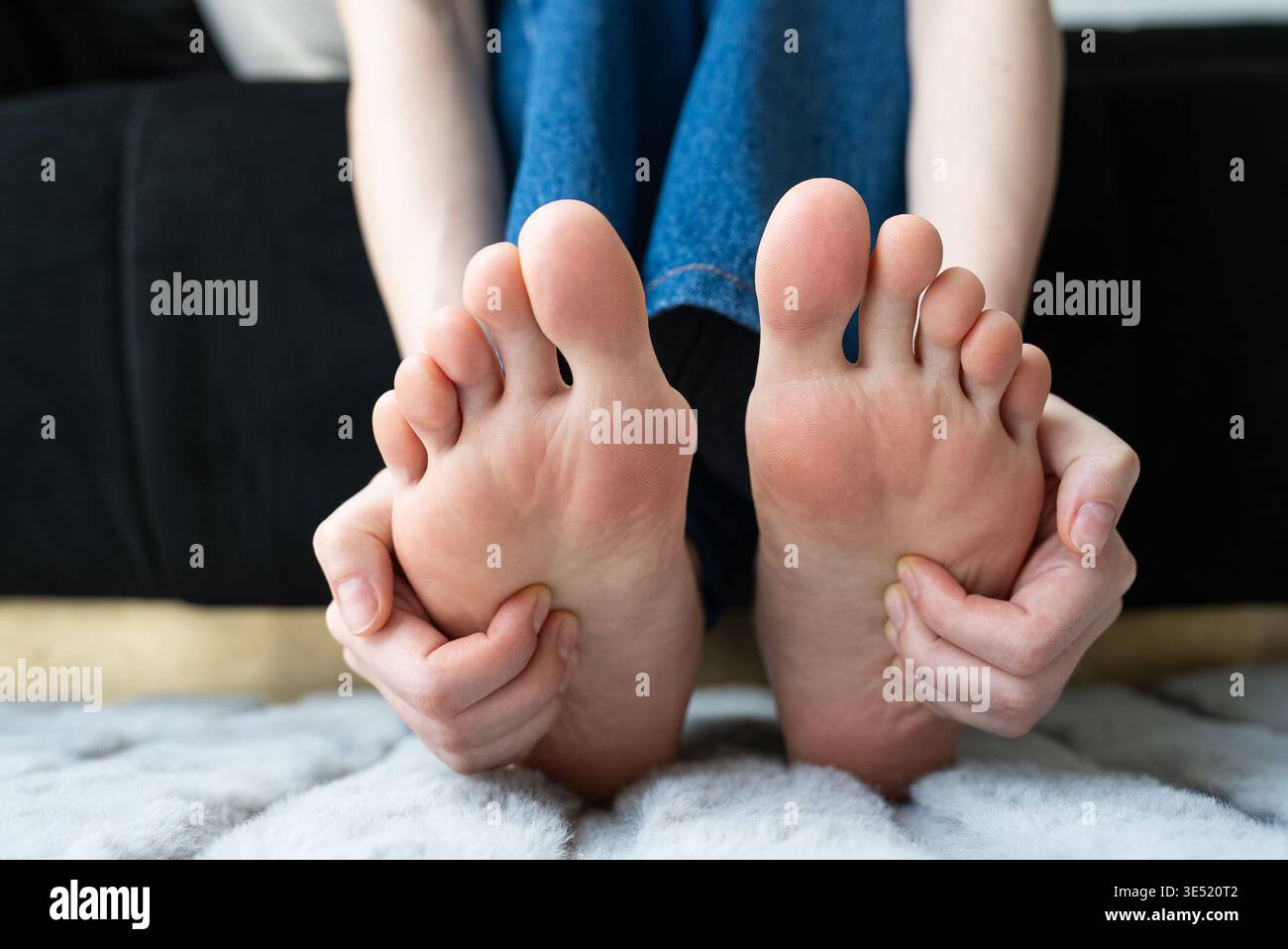 una donna si allunga e massaggia i propri piedi mentre si siede su un letto, promuovendo il relax e la cura di sé Foto Stock