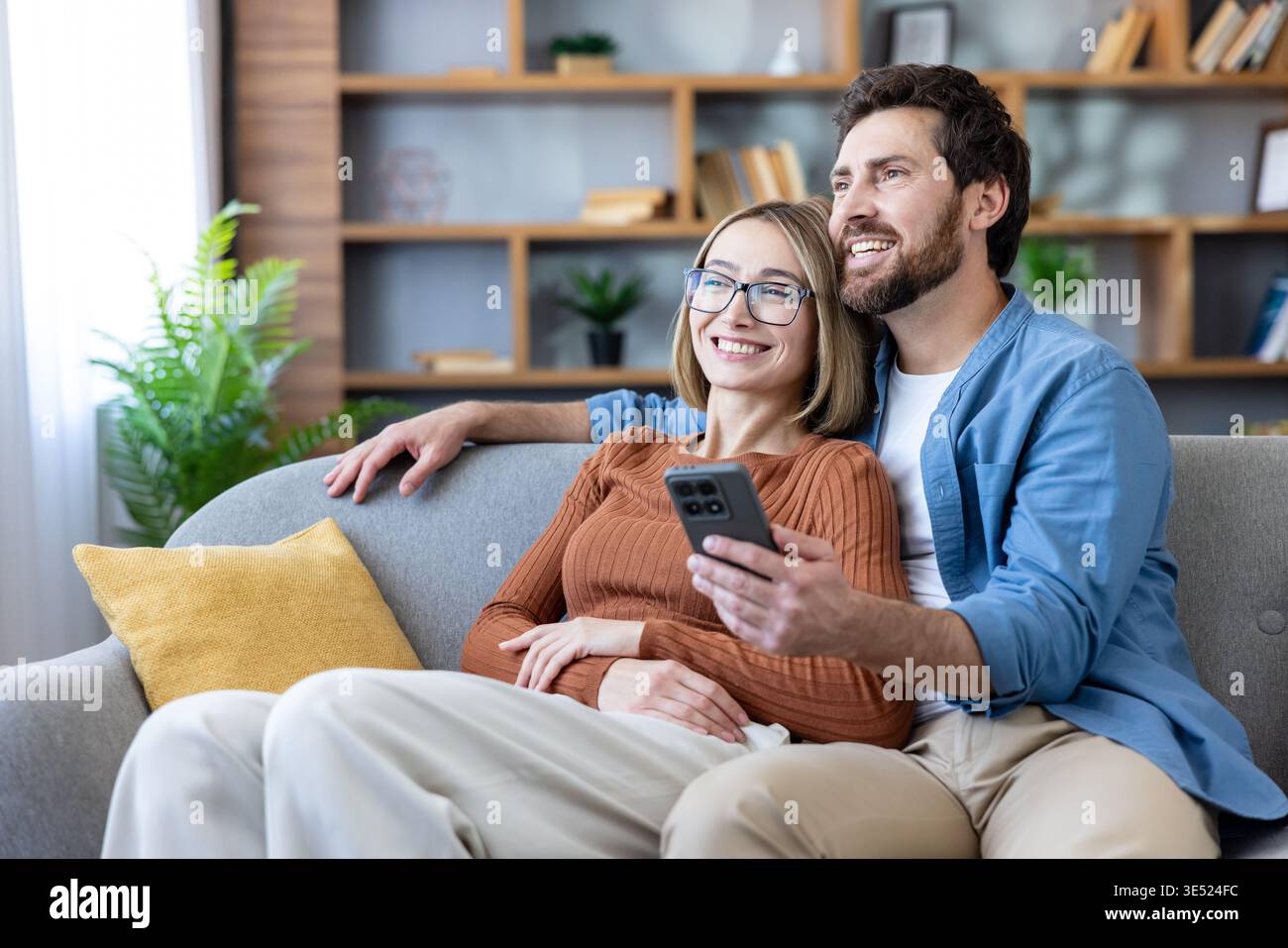 Una giovane coppia di adulti sorridente seduta insieme su un comodo divano di casa, godendosi un momento di svago guardando lo schermo di uno smartphone e condividendo un'esperienza digitale Foto Stock