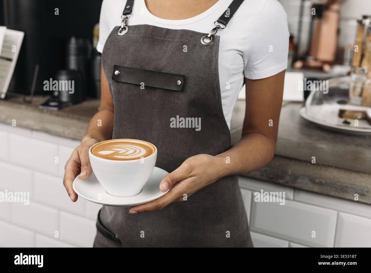 Shot ritagliato di un barista irriconoscibile che tiene in mano una tazza di caffè. Cameriera in un grembiule con un cappuccino appena preparato Foto Stock