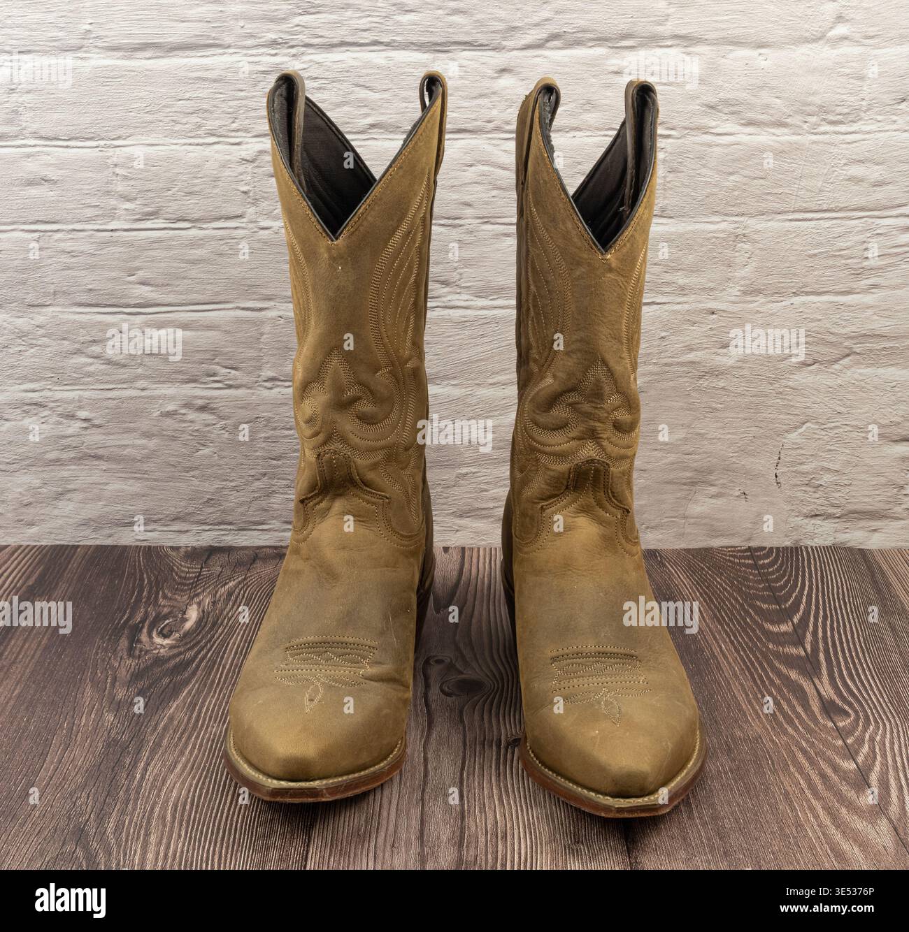 Un paio di stivali western da cowboy in pelle color marrone scuro si staglia su un pavimento di legno scuro su uno sfondo bianco in mattoni. Foto Stock