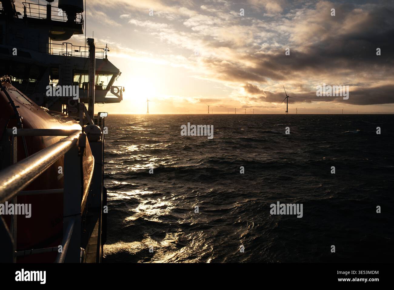 Vista dal ponte di una nave per operazioni di servizio verso un parco eolico offshore al tramonto, luce dorata sul Mare del Nord con turbine all'orizzonte. Foto Stock