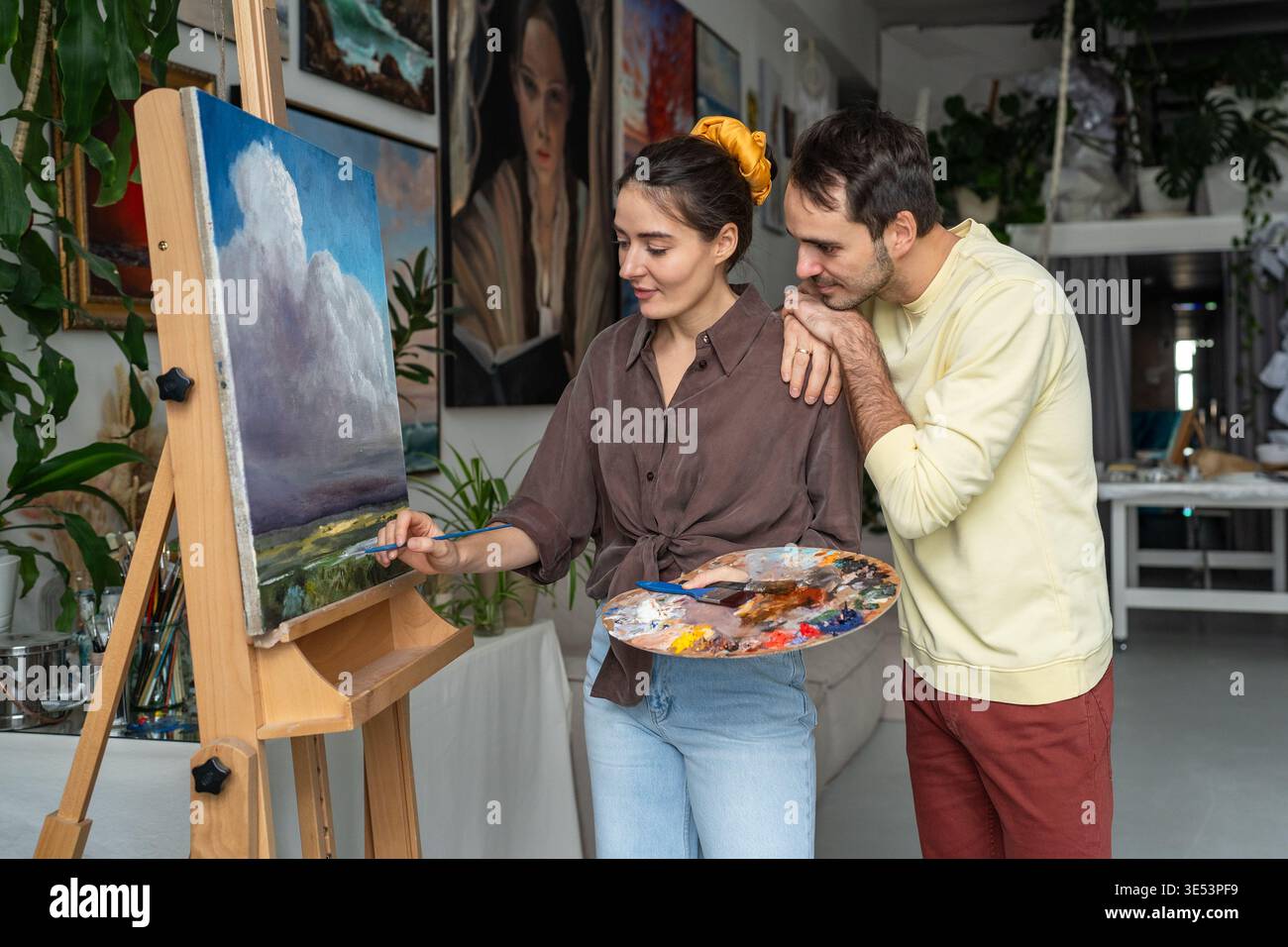 Coppia creativa che si diverte a dipingere, marito curioso che guarda la moglie disegnare un paesaggio a olio su tela. Foto Stock