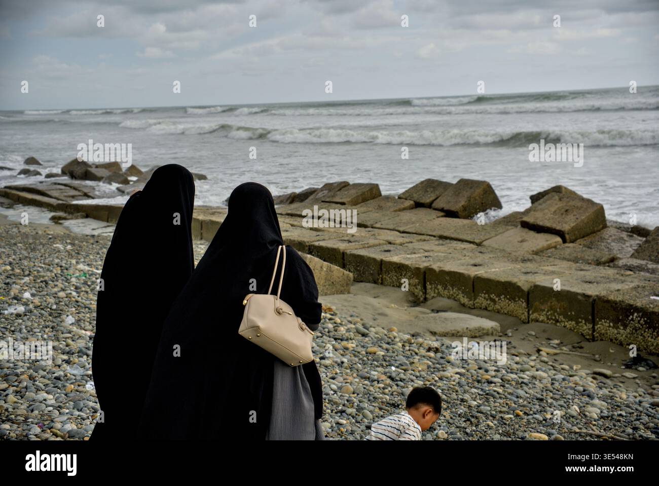 Madre e figlia camminano sulla riva con le onde che si infrangono sulla costa di Breakwater Foto Stock