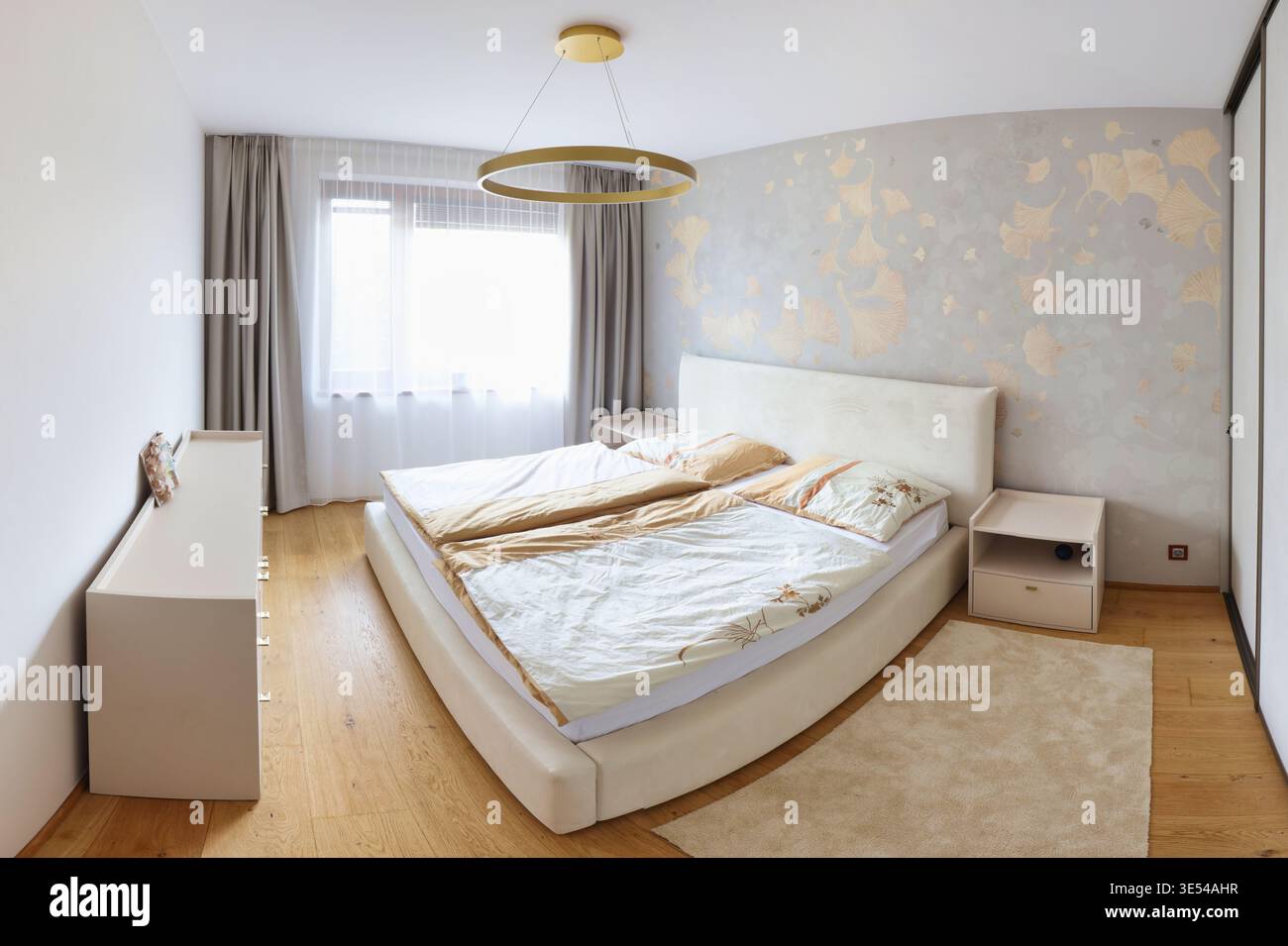 Interni contemporanei della camera da letto con pannelli in legno, letto beige, poltrona, luci a sospensione, tende morbide, e pavimenti in parquet a spina di pesce. Foto Stock
