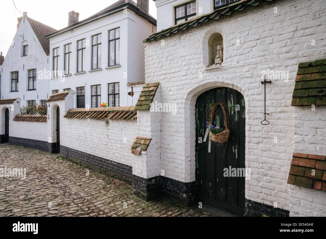 Vista di un'antica entrata nel Beghinaggio di Bruges, caratterizzata da pareti e facciate dipinte di bianco. La porta di legno scuro e' decorata con fiori, con un Foto Stock