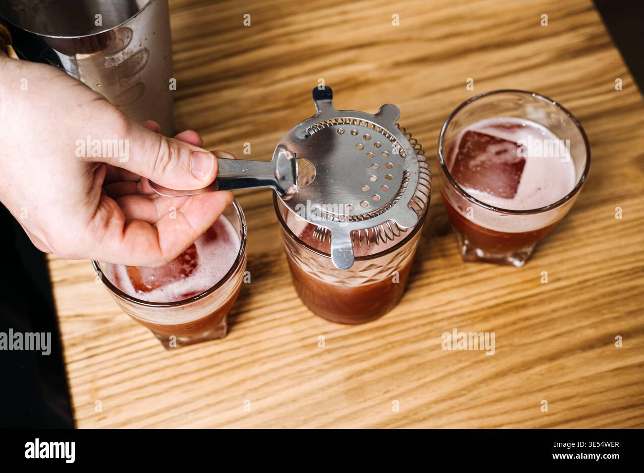 La mano tiene il filtro metallico sopra il vetro riempito con liquido rosso e ghiaccio insieme ad altre due bevande. Bere consapevole della salute, rituale senza alcol Foto Stock