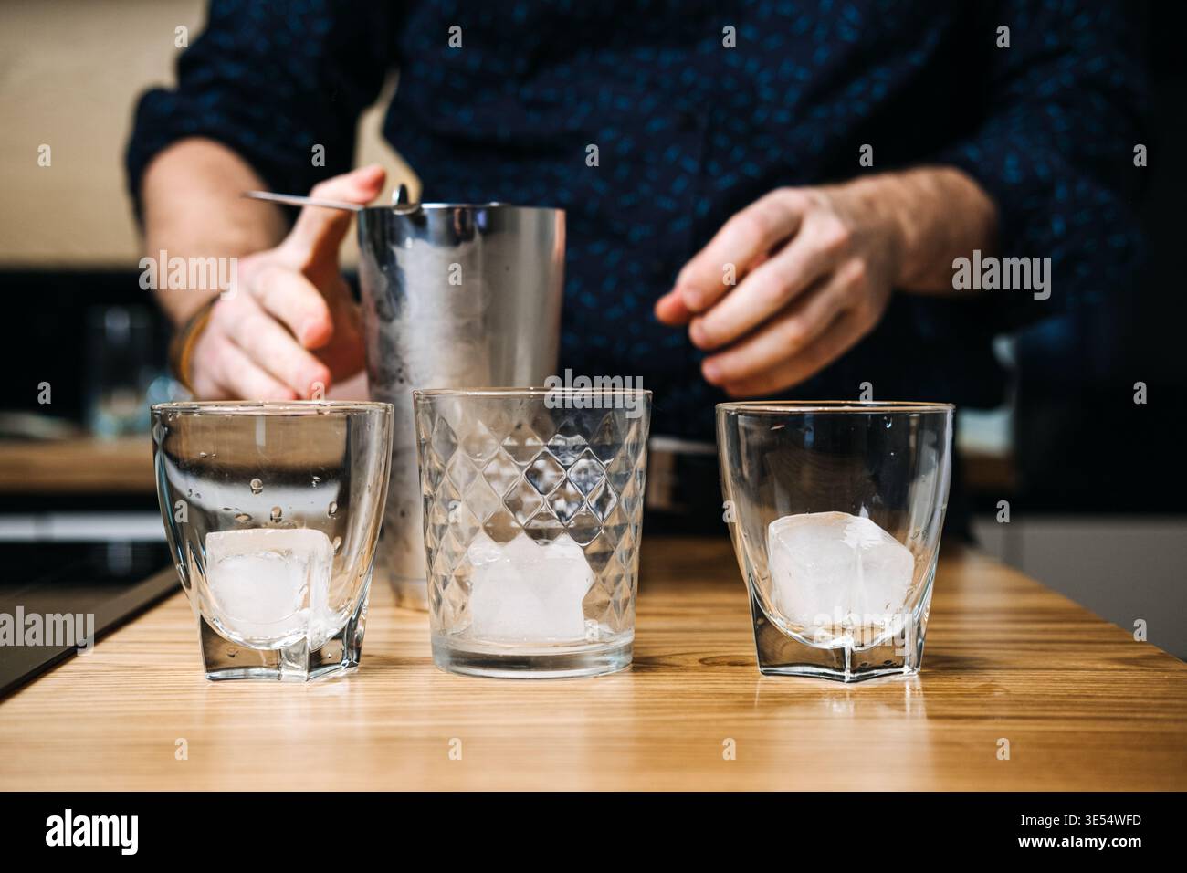 Tre bicchieri con cubetti di ghiaccio siedono sul tavolo di legno mentre la persona prepara lo shaker per cocktail sullo sfondo. Cocktail fai-da-te, bevande a casa, fine settimana Foto Stock