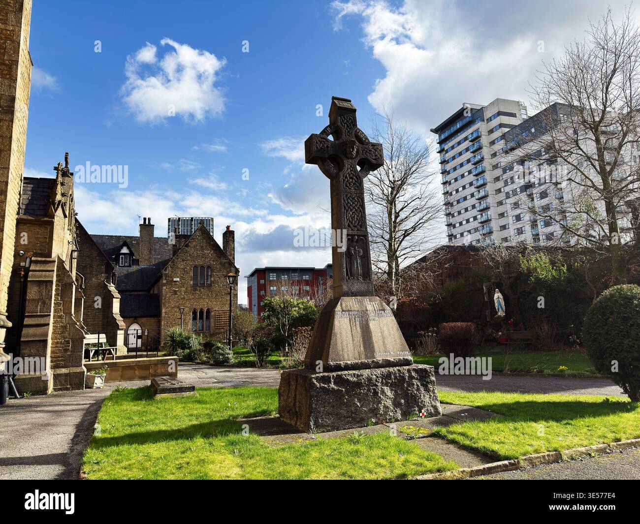 Manchester, Regno Unito - 22 febbraio 2026: Monumento ornato a croce celtica in un giardino di sagrato incorniciato da edifici storici in pietra e appartamenti moderni. Orat Foto Stock