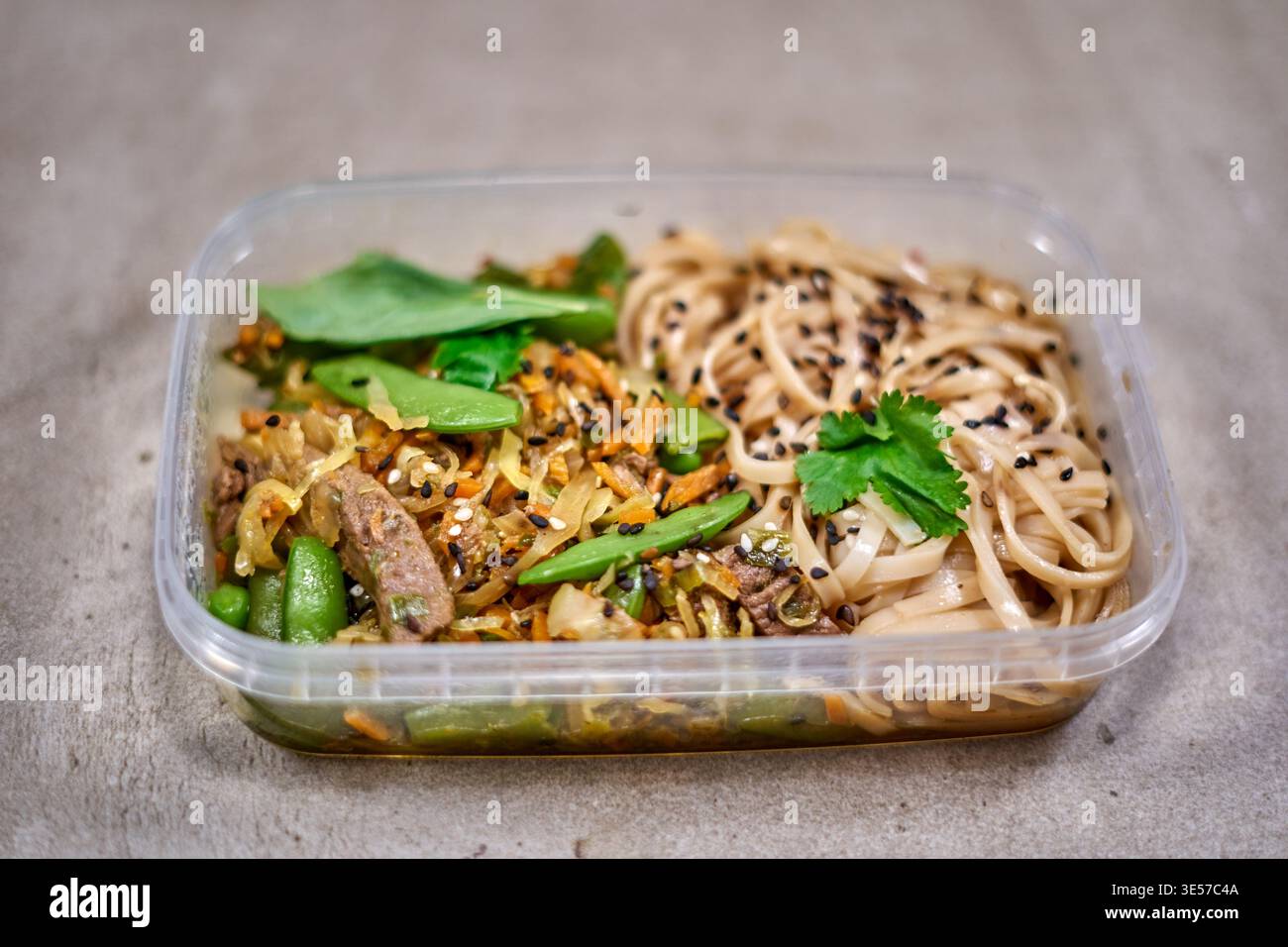 Un contenitore rettangolare in plastica trasparente per la preparazione dei pasti riempito con un piatto di ispirazione asiatica si trova su una superficie ruvida grigia. Il container è Foto Stock