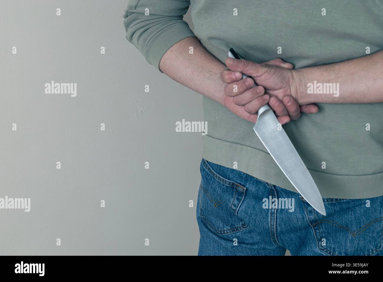 Primo piano di un uomo che tiene un coltello da cucina dietro la schiena, che suggerisce pericolo, minaccia o intento nascosto. Concetto di violenza, crimine, segretezza o potenziale Foto Stock