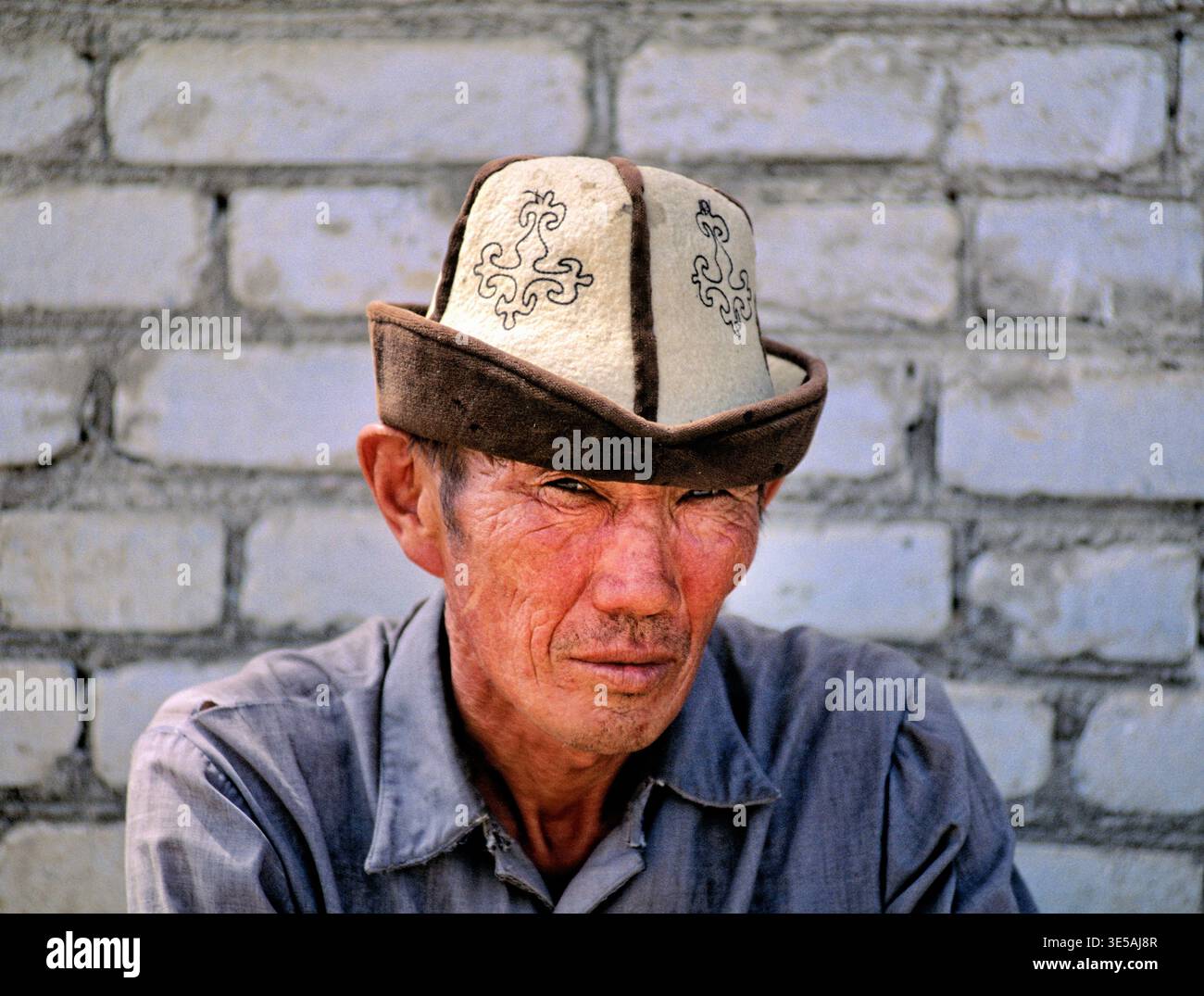 Un uomo che indossa un cappello tradizionale si siede tranquillamente contro un muro di mattoni a Bukhara, Uzbekistan. La sua espressione mostra un pensiero profondo e una riflessione. Foto Stock