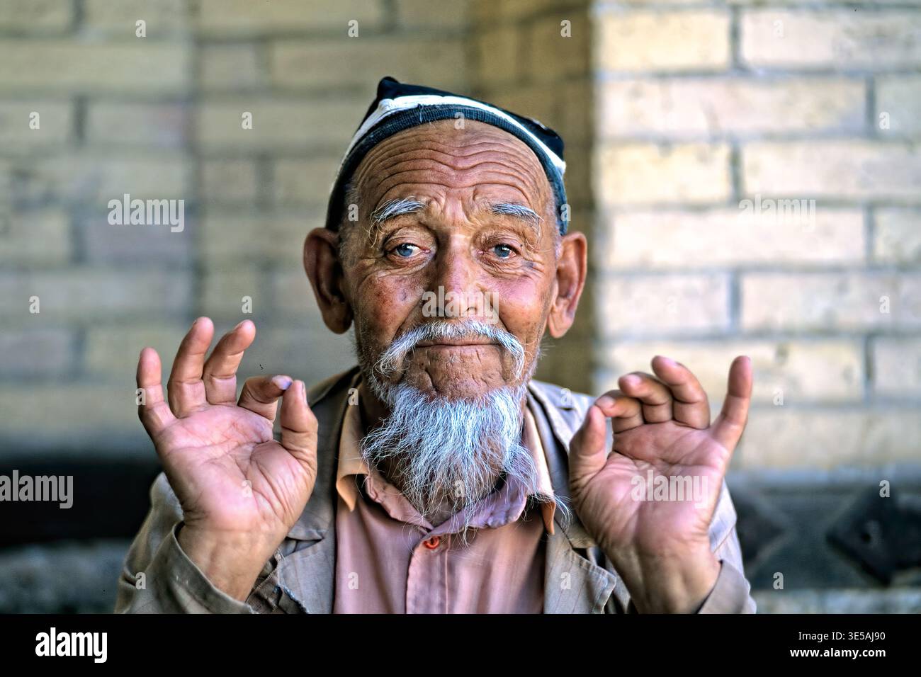Un uomo anziano siede a Khiva, in Uzbekistan, e mostra gesti delle mani con le mani alzate. Sorride calorosamente ai passanti. Foto Stock