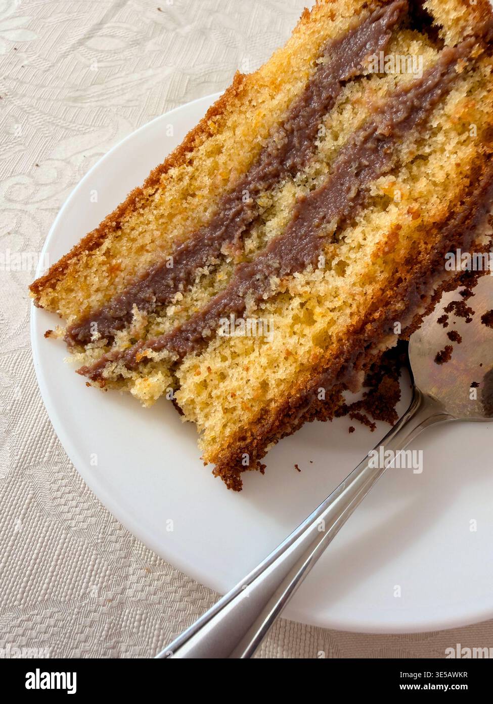 Primo piano di un pezzo di torta in spugna senza glutine e senza lattosio multistrato con crema di cioccolato su un piatto bianco con cucchiaino - Immagine stock catturata con smartphone