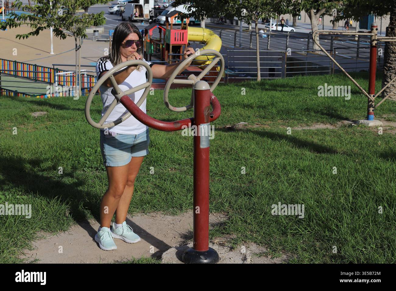 Donna che si allena su attrezzature fitness all'aperto in un parco con strutture parco giochi e vegetazione visibile sullo sfondo Foto Stock