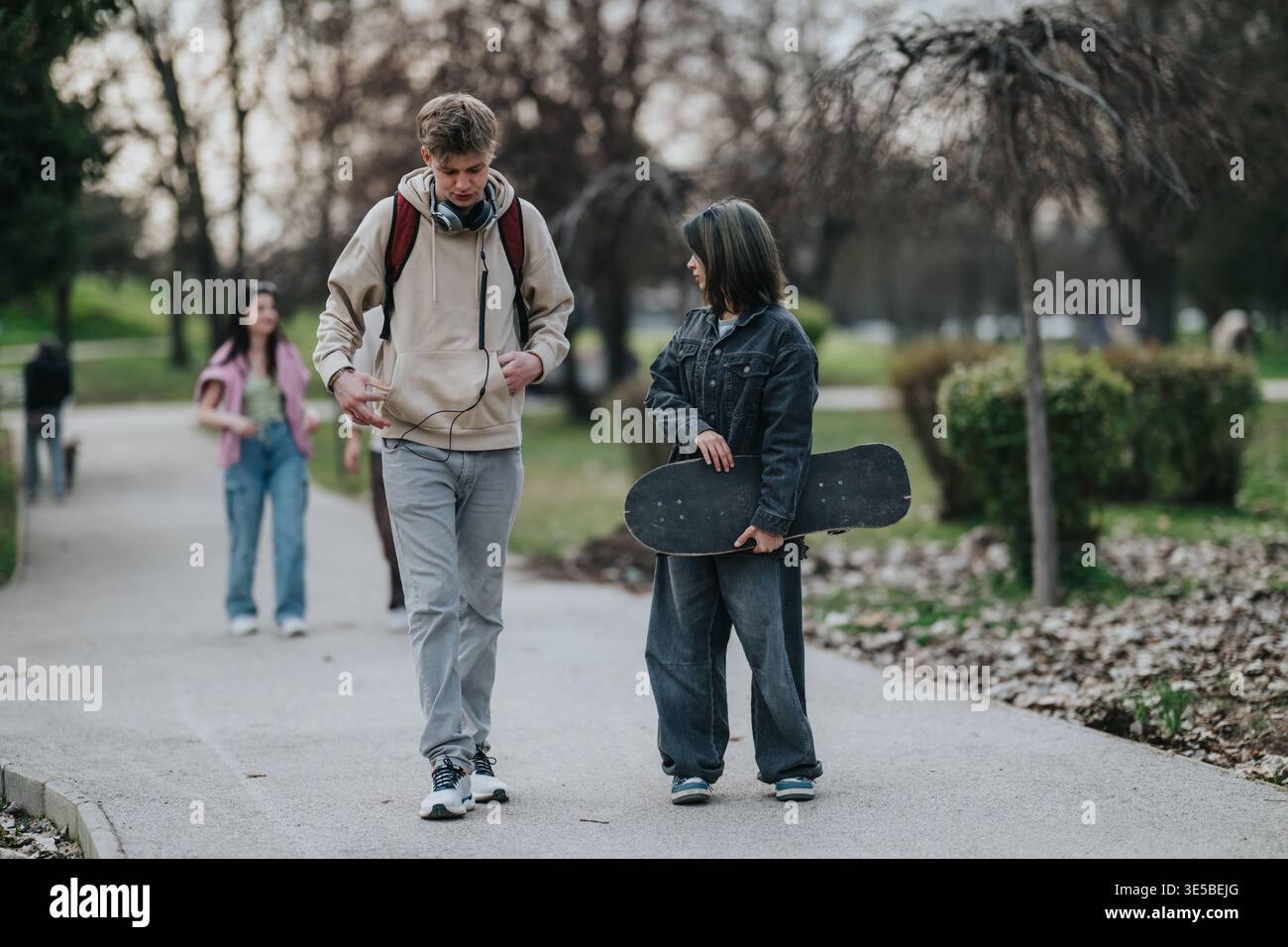 Due adolescenti con uno skateboard che camminano e parlano in un parco con amici informali che escono Foto Stock