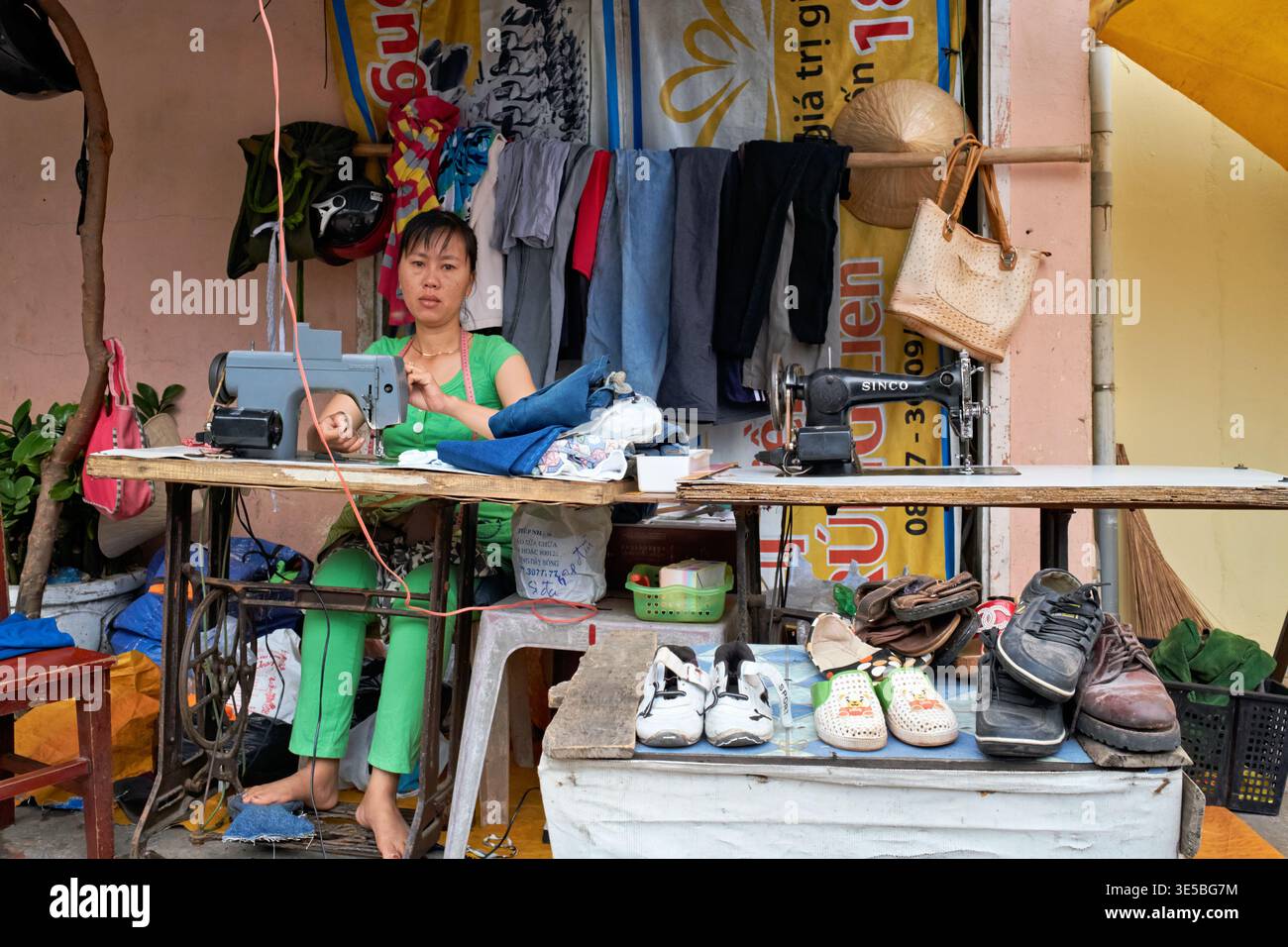 Una donna cuce vestiti al suo stand a Phu Quoc, in Vietnam. È concentrata sul suo lavoro mentre è circondata da vestiti e scarpe. Foto Stock