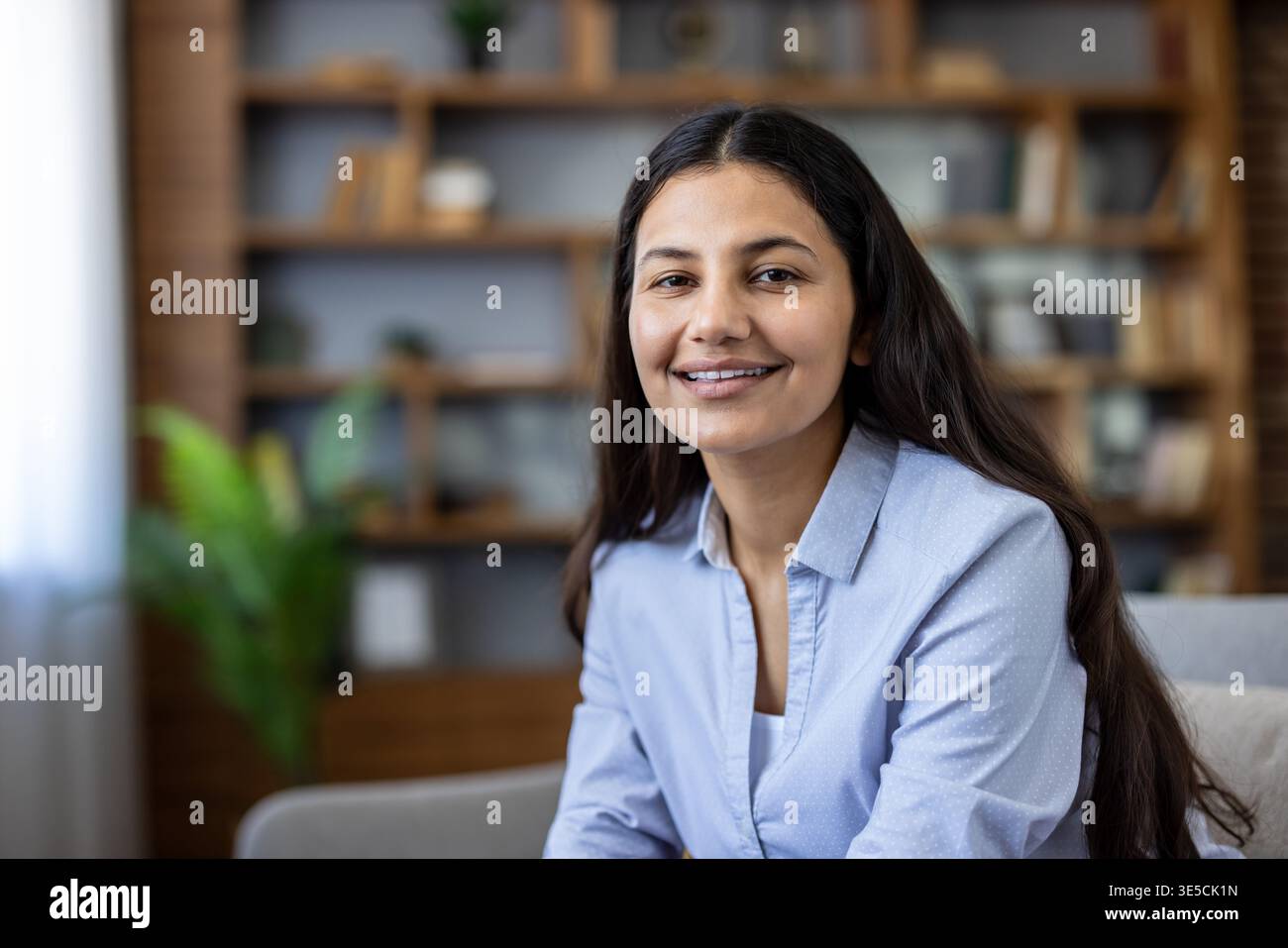Giovane donna indiana che sorride con fiducia su un comodo divano in una casa moderna, rilassata e accessibile con uno sfondo sfocato da libreria, ideale per lo stile di vita e concetti di lavoro a distanza Foto Stock