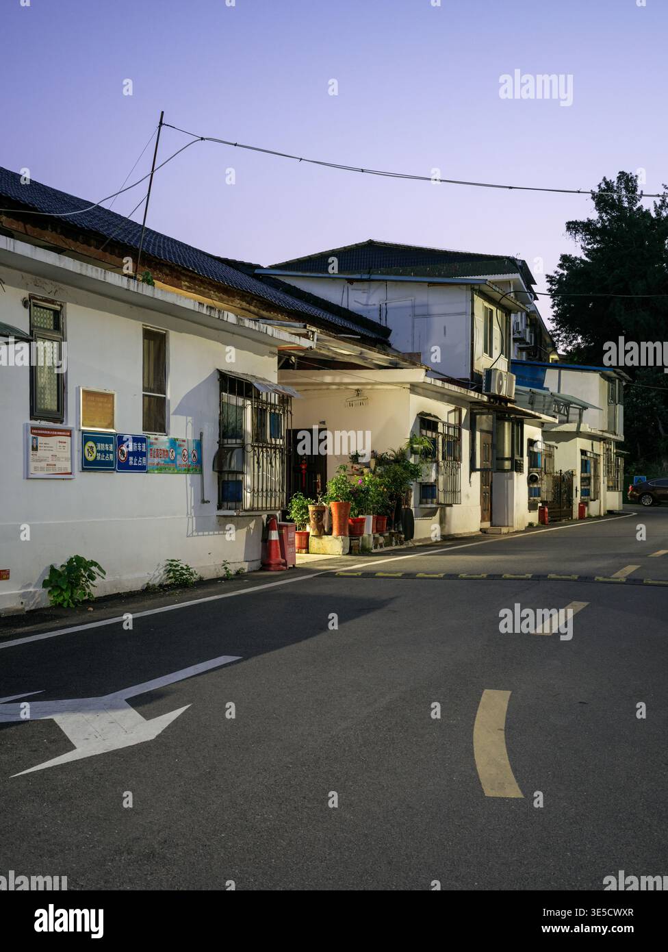 Wuhan, Cina - 16 giugno 2025: Strada pavimentata con frecce bianche e corsie gialle che conducono davanti a vecchi edifici residenziali a due piani con la facciata bianca Foto Stock