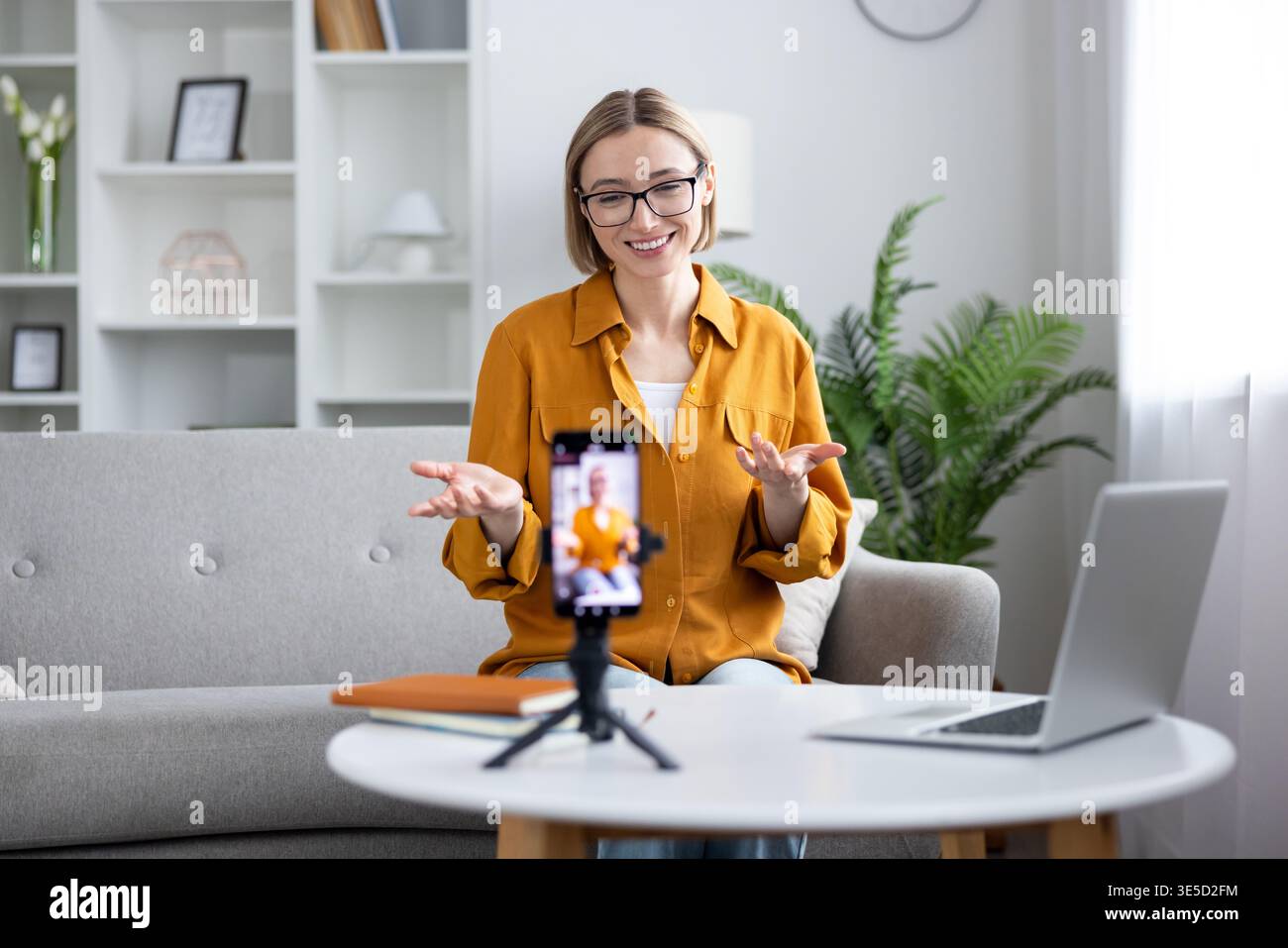 Donna che registra un video blog o un corso online a casa, siede su un divano e utilizza uno smartphone su un cavalletto, creando nuovi contenuti digitali per il suo pubblico Foto Stock