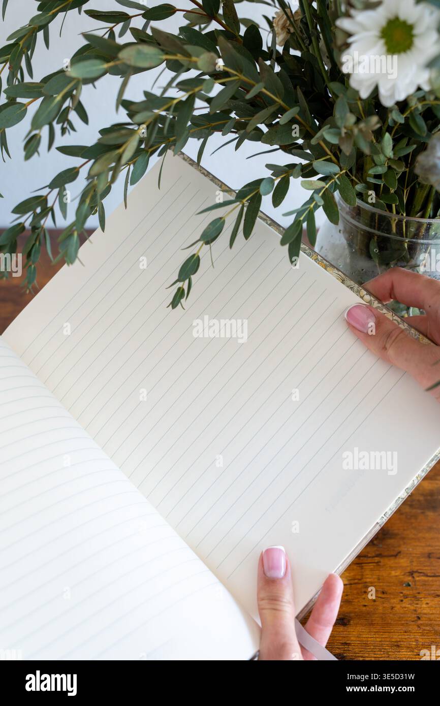 Primo piano di mani femminili che tengono in mano un notebook bianco, circondato da fiori verdi, stile di vita e concetto di produttività foto verticale Foto Stock