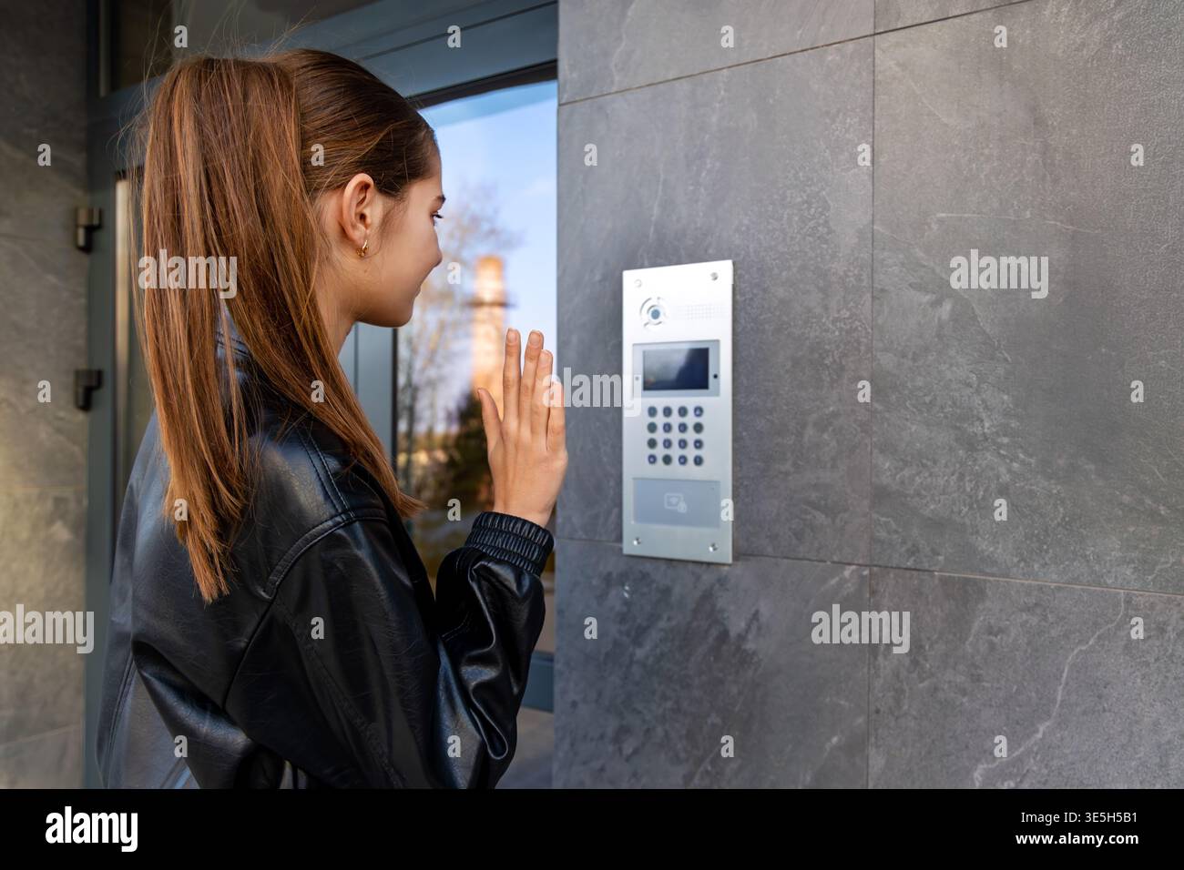 Giovane donna che usa l'Intercom Appartamento all'esterno. Una giovane donna con una giacca in pelle nera utilizza un pannello interfono all'aperto per un moderno ingresso di un appartamento Foto Stock