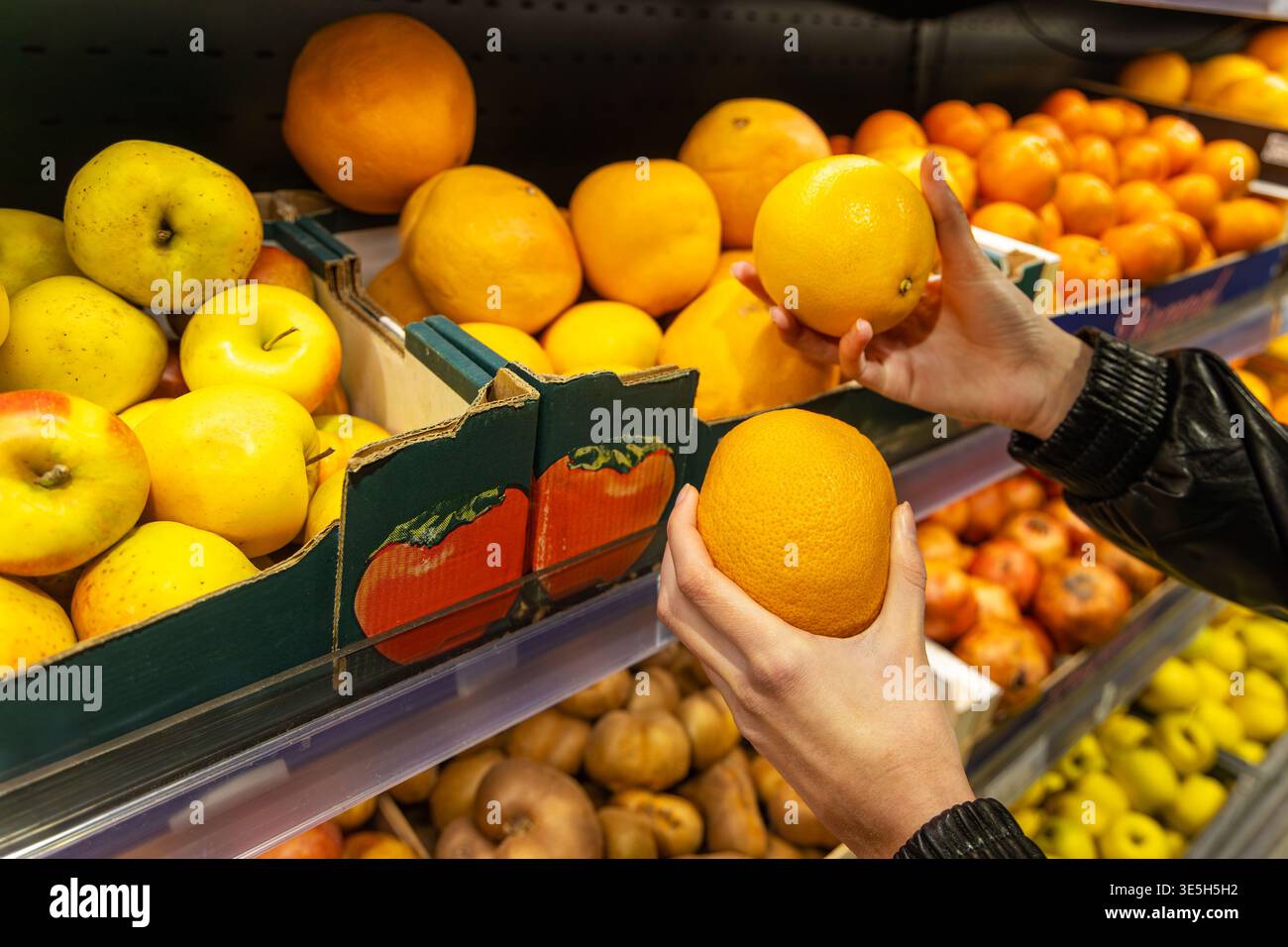 Mani che scelgono le arance nel corridoio della frutta del supermercato. Primo piano di mani che selezionano arance fresche in un corridoio di frutta del supermercato, circondato da mele e. Foto Stock