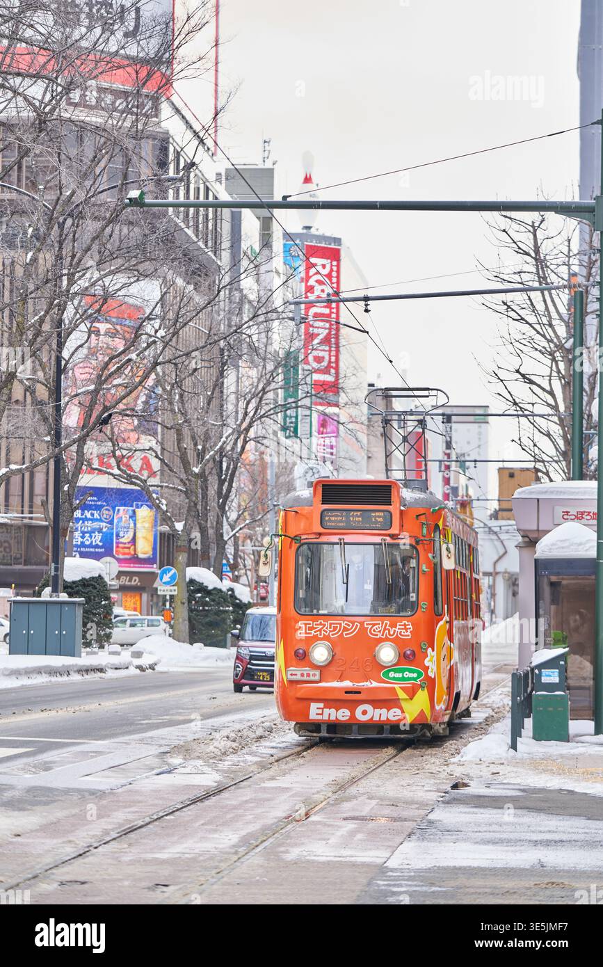 Una pittoresca scena stradale invernale a Sapporo, Hokkaido. Un tram d'epoca arancione corre lungo le piste innevate, creando un m nostalgico e romantico Foto Stock