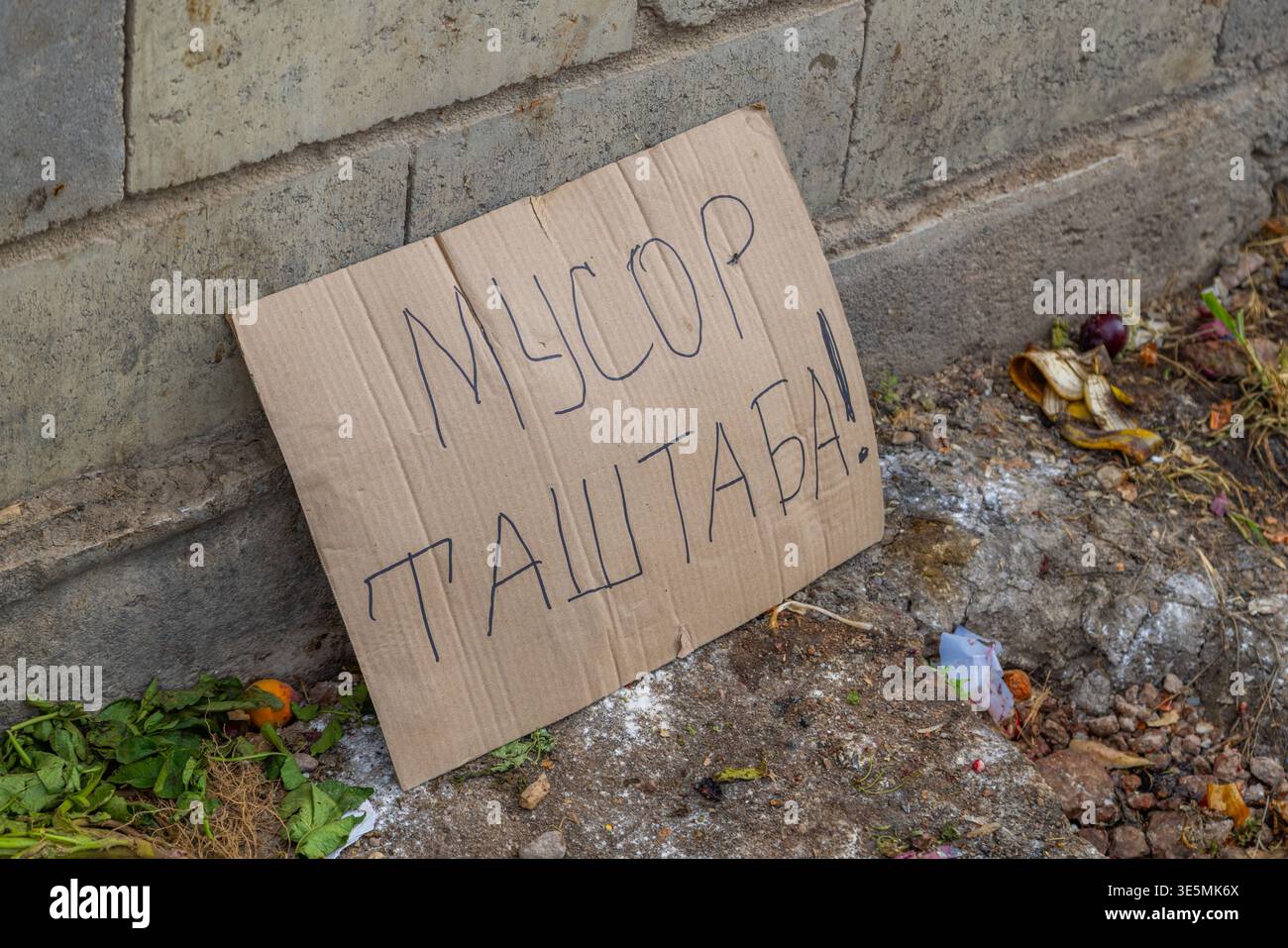Cartello di cartone indicante i rifiuti alimentari sulle strade urbane scritto in Kirghizistan che significa non gettare rifiuti. I rifiuti circostanti e le pareti di cemento raffigurano il decadimento e l'abbandono urbano. Foto Stock