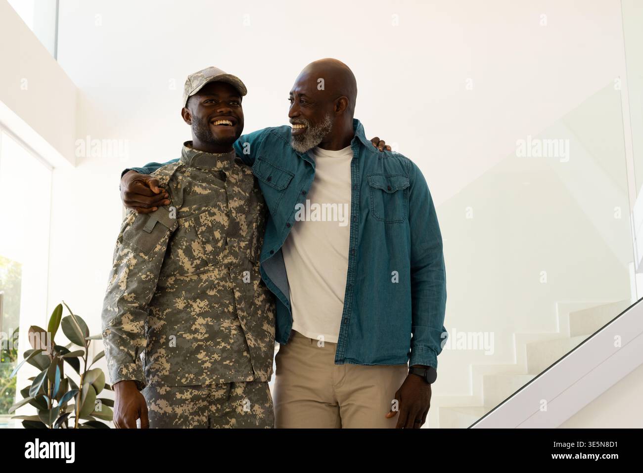 Padre e figlio afroamericani sorridenti in piedi nel foyer, con uniforme mimetica e cappello Foto Stock