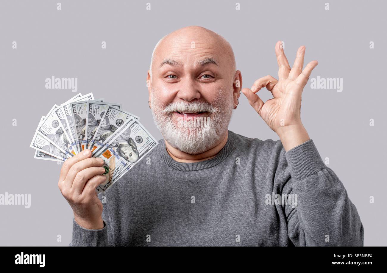 Uomo anziano che tiene banconote in dollari con le dita dei cartelli, gesti amichevoli sorridenti simbolo eccellente isolato su sfondo grigio. Uomo in attesa Foto Stock