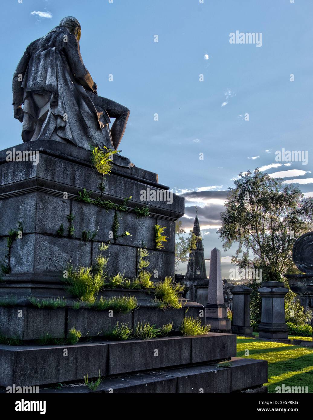 Una suggestiva necropoli al crepuscolo, caratterizzata da una statua intemprata in cima a un monumento in pietra, erba e felci sovrastate, tombe storiche e un umore Foto Stock