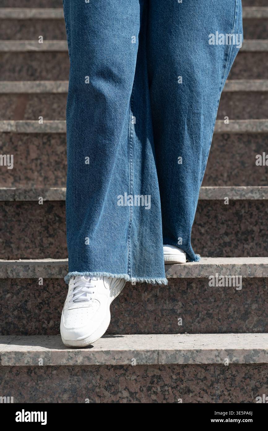 Vista ad angolo basso della persona che indossa jeans blu a gamba larga e sneakers bianche che camminano giù per le scale di granito. Foto Stock