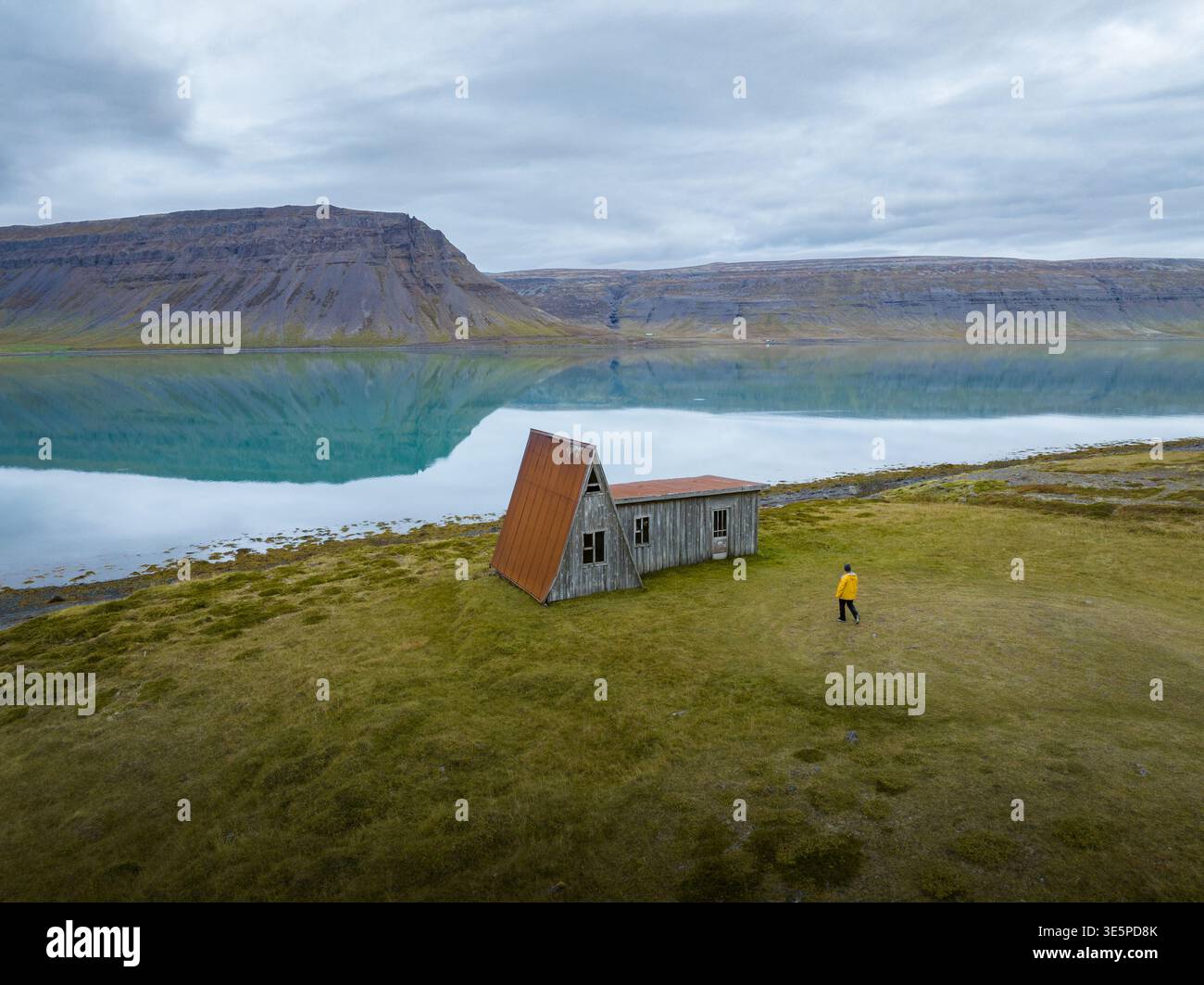Vista aerea di una figura solitaria in giallo in piedi vicino a una casa abbandonata con un tetto rosso, riflessa nelle acque ancora fiordi, Westfjords, Islanda. Foto Stock
