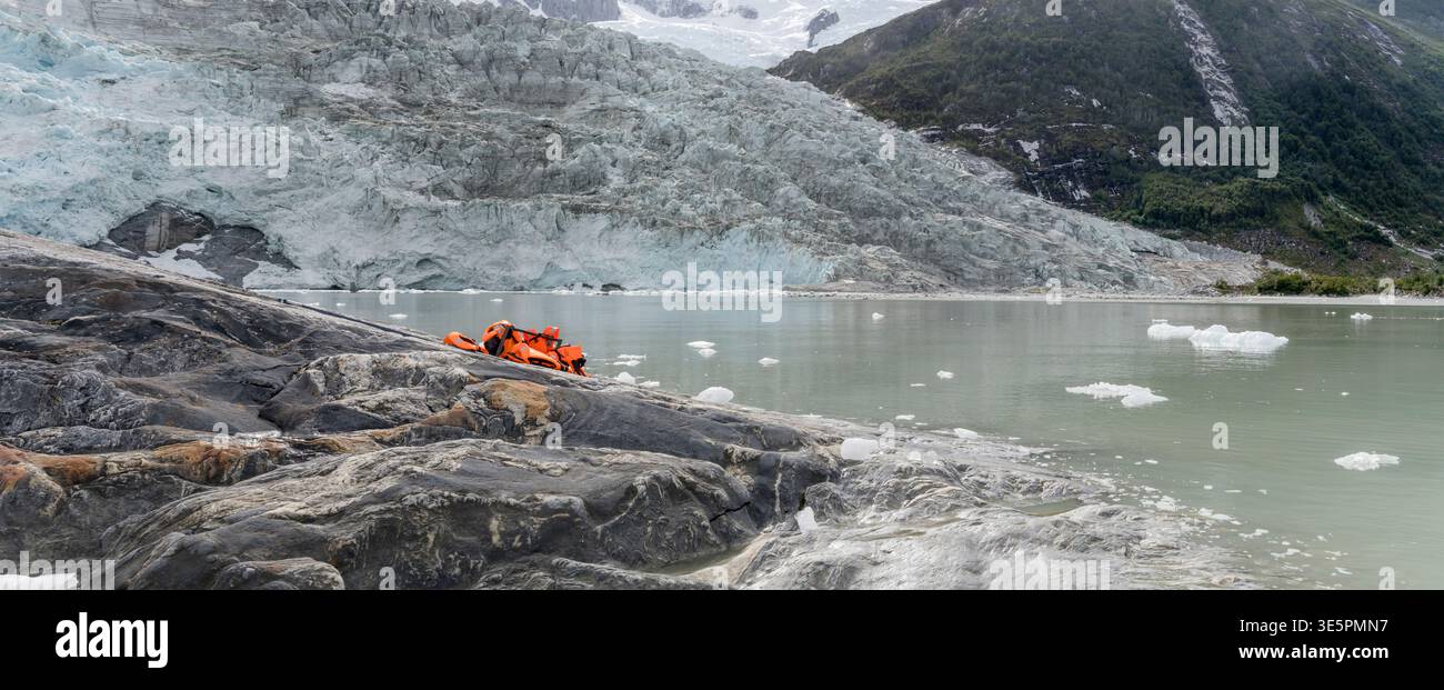 Paesaggio con giubbotti di salvataggio rossi sulla costa rocciosa e parete di ghiaccio sul fronte orientale del ghiacciaio sull'acqua del fiordo, girato con la luce estiva al fiordo di Pia, Cile Foto Stock