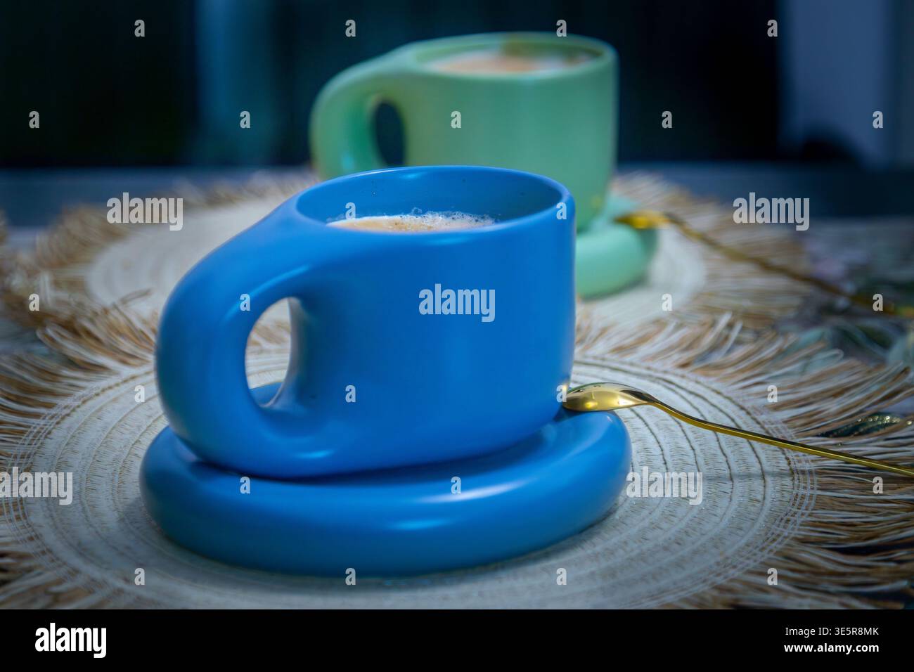 Latte servito in una moderna tazza blu disegnata posta su una placchetta rustica intrecciata con una seconda tazza leggermente sfocata sullo sfondo, un'accogliente scena di natura morta Foto Stock