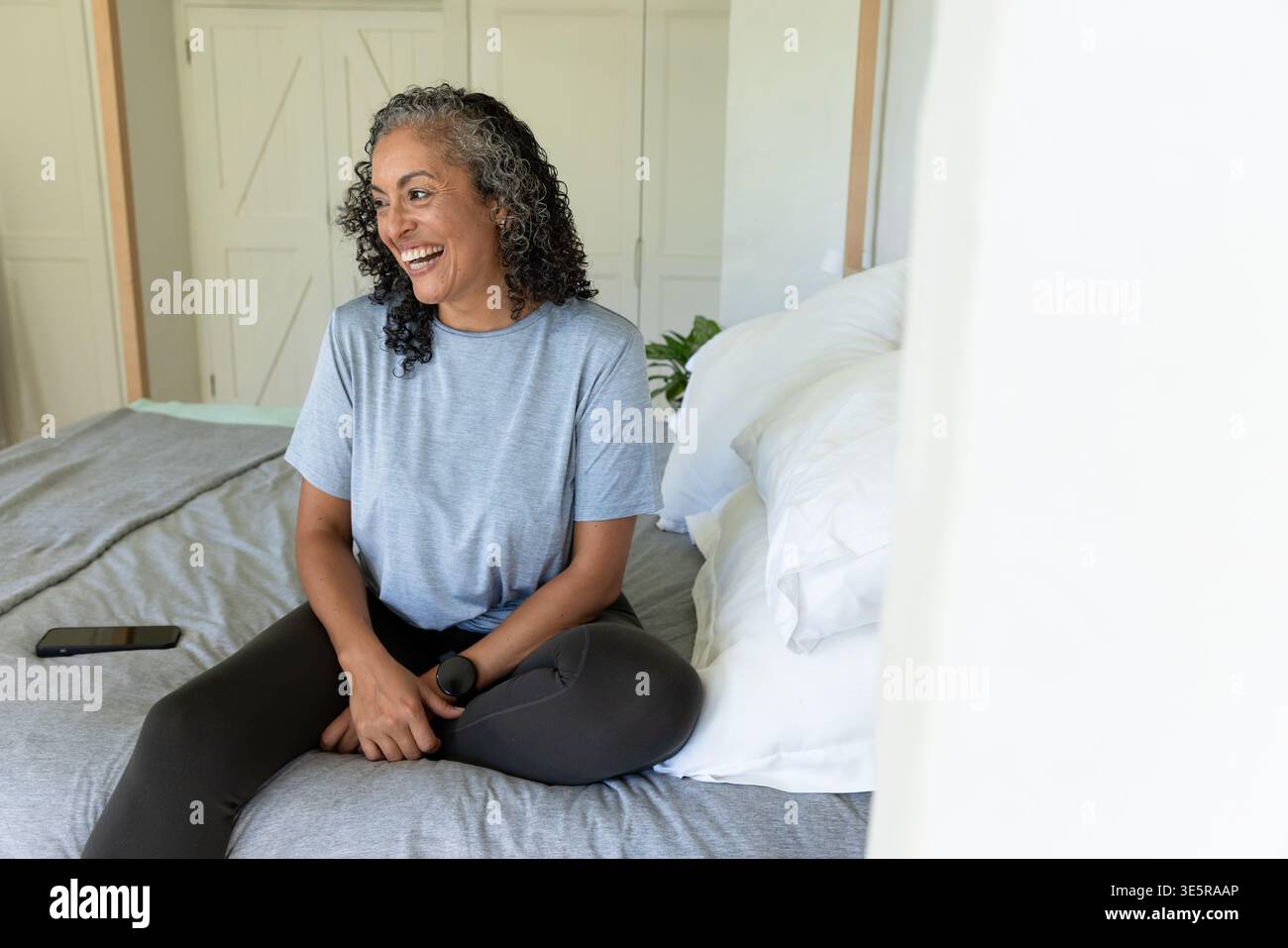 Donna afroamericana seduto sul letto con tee grigie e leggings sorridenti sullo smartphone Foto Stock