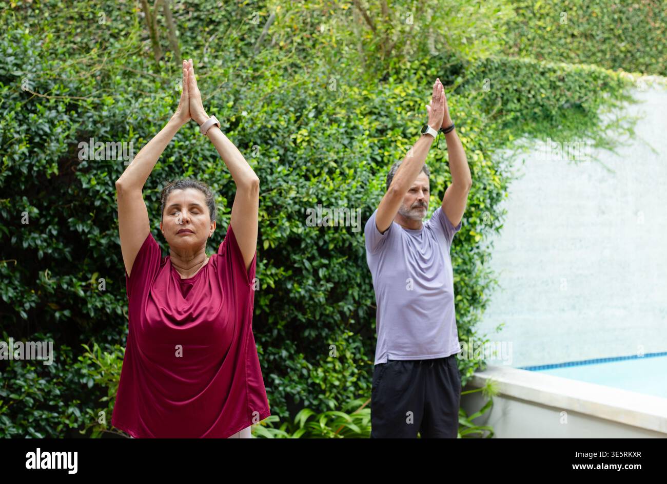 Coppia matura che pratica yoga in piedi in giardino accanto alla piscina con orologi da polso visibili Foto Stock