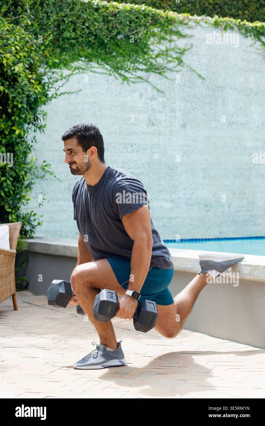 Uomo che fa un affondo a una gamba sul patio che regge i manubri indossando pantaloncini a parete bassa e smartwatch Foto Stock